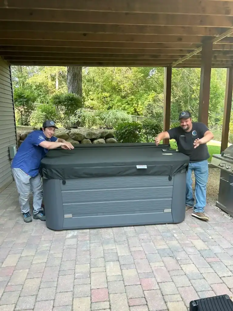 Two men pose and smile next to a covered outdoor hot tub on a stone patio under a wooden pergola, with trees and landscaping behind them, giving an inviting glimpse into the spirit of our "about us" story.