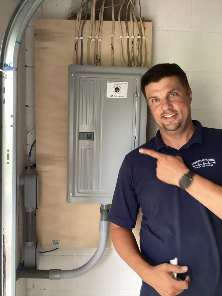 A man wearing a navy blue shirt stands indoors, smiling and pointing at a mounted electrical panel with multiple wires above it, showcasing his expertise—learn more on our About Us page.