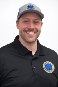 Meet Our Family! Man wearing a black collared shirt and gray cap with matching logos, smiling at the camera against a plain white background.