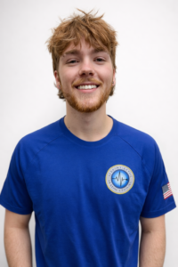 Meet Our Family! A young man with light brown hair and a beard, wearing a blue T-shirt with a circular logo and an American flag patch, stands against a plain white background, smiling at the camera.