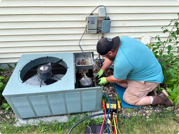 A technician kneels beside a house, servicing an outdoor air conditioning unit with tools and gauges connected.