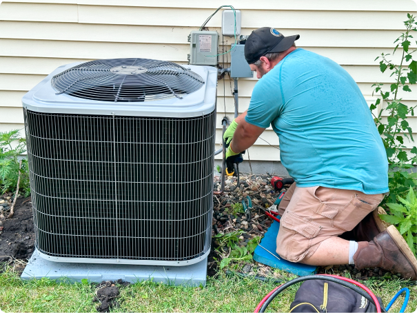 A technician in a blue shirt and gloves is repairing or servicing an outdoor air conditioning unit next to a house.